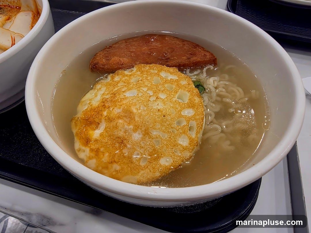 A close-up shot of a bowl of noodle soup. The soup contains instant noodles, a fried egg, and a slice of pan-fried meat. Another bowl with what appears to be a spicy soup with white slices is visible to the left.