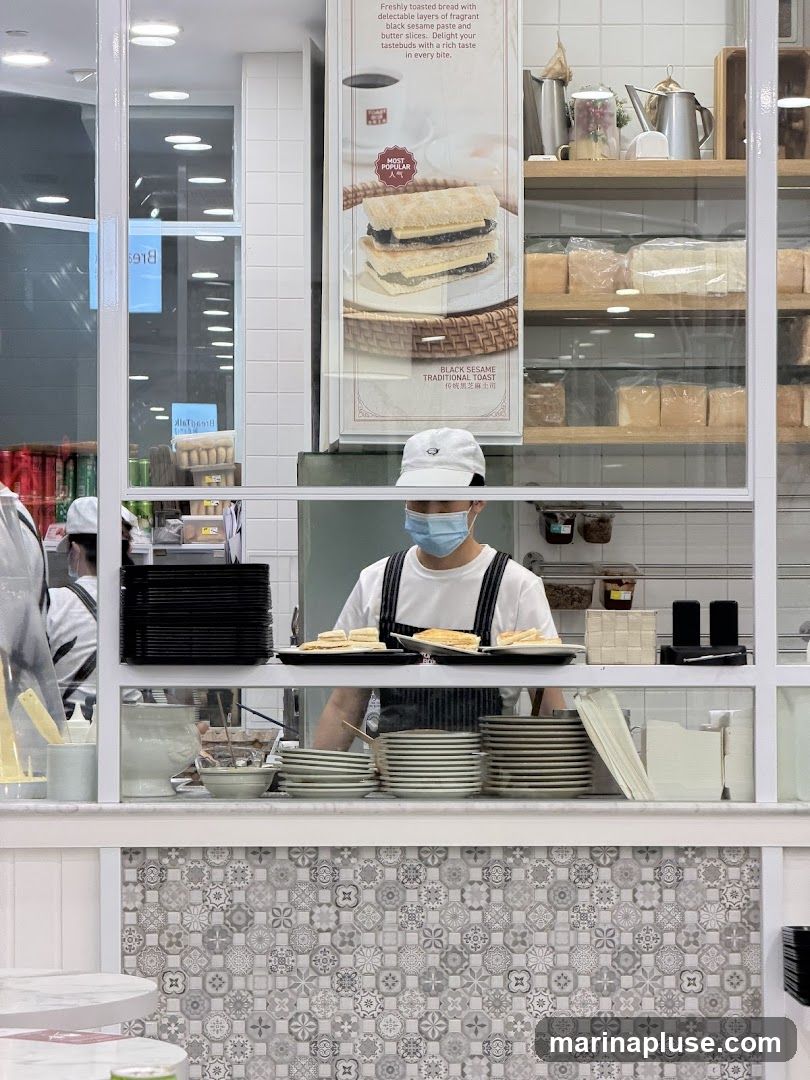 A person wearing a mask and a white cap is preparing toast behind a glass partition. Stacks of plates and bread are visible. A poster advertises "Black Sesame Traditional Toast" as "Most Popular."