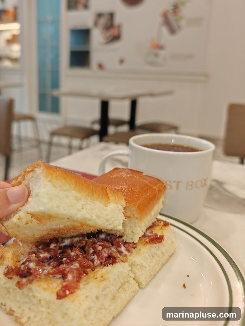 A close-up shot of a person holding a half-eaten toast with a creamy filling, served on a white plate with a green rim. A white mug of coffee sits in the background, with the words "POST BOX" visible on its side. The setting appears to be a cafe with tables and chairs visible in the blurred background, along with some wall art.