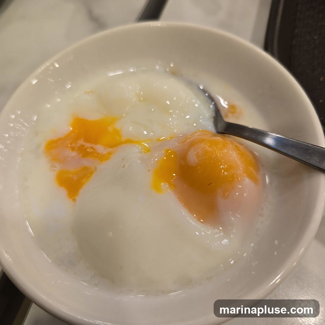 A close-up shot of two soft-boiled eggs in a white bowl, with a spoon resting in the bowl. The egg yolks are runny and bright orange, contrasting with the white of the egg whites and the surrounding liquid.