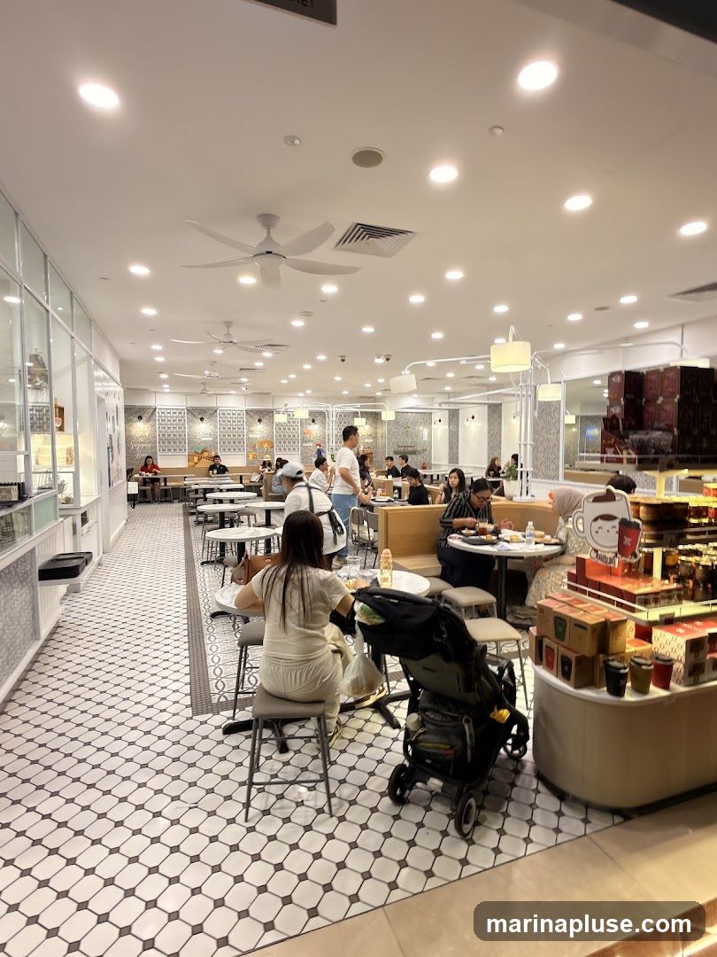 The image shows the interior of a brightly lit cafe with white tiled floors and white walls. Several tables are occupied by diners. A woman in a white outfit is seated at a table in the foreground, with a stroller next to her. In the background, more tables and patrons are visible, along with ceiling fans and modern light fixtures. A display counter with boxes of products is on the right side of the image.