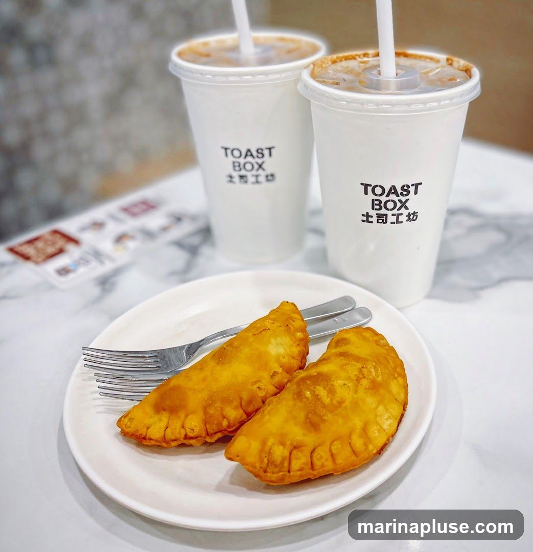 Two white cups with the "Toast Box" logo and Chinese characters are in the background. In the foreground, a white plate holds two golden-brown pastries and a fork. A blurred menu is visible on the left.