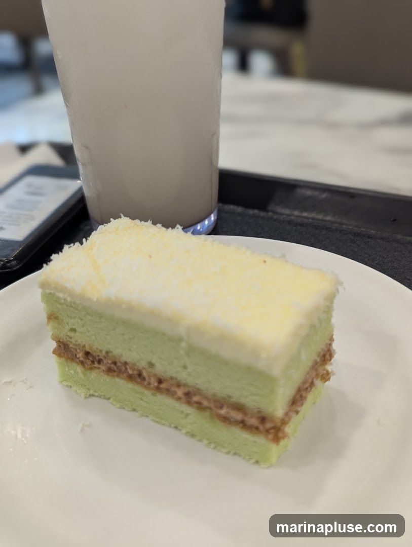 A slice of green cake with white frosting and a layer of filling is served on a white plate. A tall glass of a light-colored beverage, possibly a milkshake or smoothie, is in the background. A dark tray holds the plate and glass, and a blurred electronic device is visible to the left. The setting appears to be a cafe or restaurant.