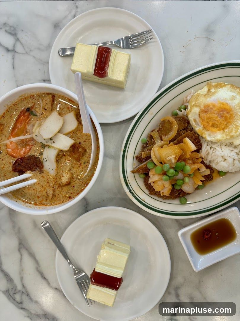 A top-down view of a marble table with two slices of layered cake with red jam, a bowl of laksa, a plate of rice with a fried egg and stir-fried ingredients, and a small dish of sauce.