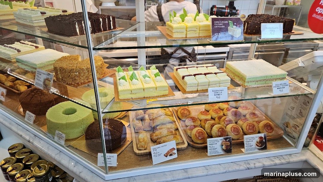 A display case filled with various cakes, pastries, and jars of preserves. Visible items include chocolate cake, corn custard tea cake, raspberry vanilla cake, gula melaka coconut cheesecake, bak kwa pastry, traditional salty tau sar piah, and traditional sweet tau sar piah. Jars of preserves are also visible at the bottom left.