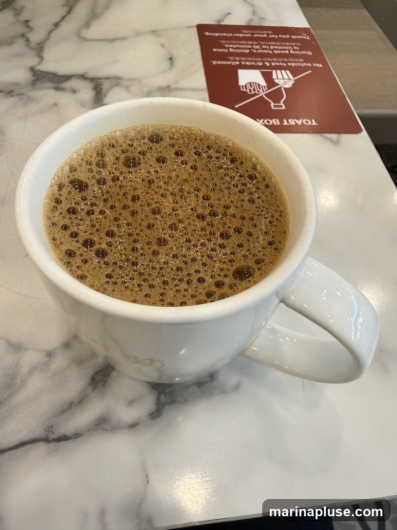 A white mug filled with a frothy brown beverage, likely coffee, sits on a marble-patterned table. To the right of the mug is a red sign with white text and graphics. The sign reads "TOAST BOX" and states that outside food and drinks are not allowed, with dining time limited to 30 minutes during peak hours.