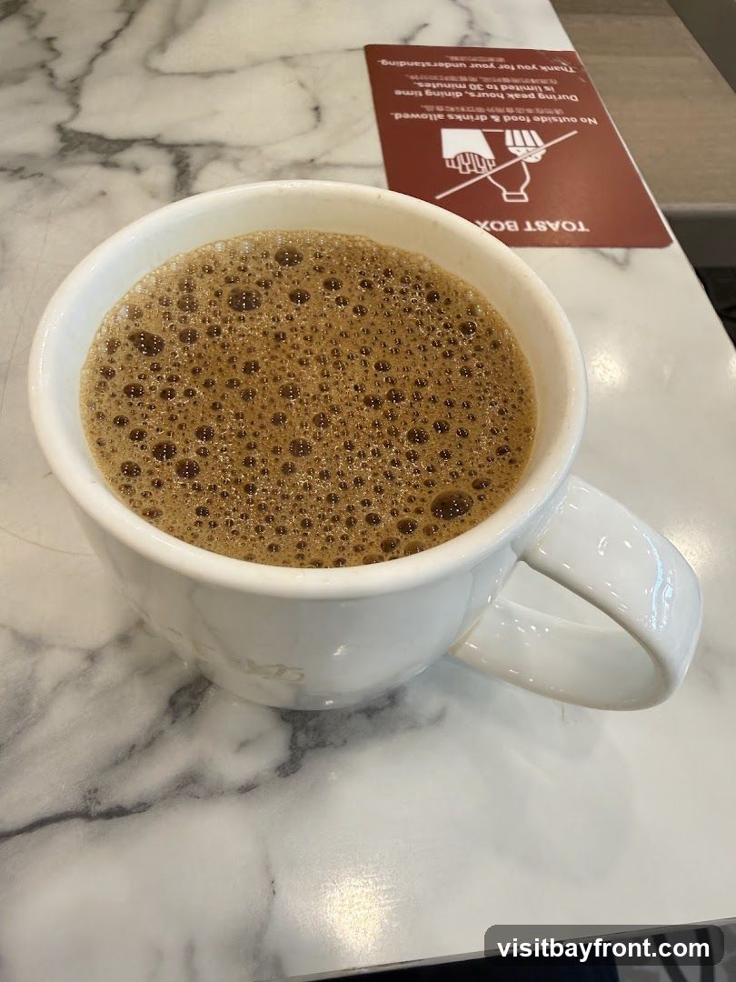 A white mug filled with a frothy brown beverage, likely coffee, sits on a marble-patterned table. To the right of the mug is a red sign with white text and graphics. The sign reads "TOAST BOX" and states that outside food and drinks are not allowed, with dining time limited to 30 minutes during peak hours.