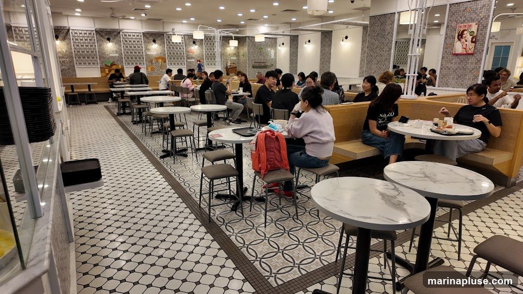 A busy restaurant interior with many patrons seated at tables. The decor features patterned tiles on the floor and walls, with modern lighting fixtures. Tables are round and oval, with chairs and banquette seating. Some patrons are eating, while others are conversing. A poster with a woman's image is visible on the wall.