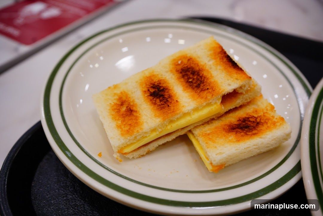 A close-up shot of two halves of a grilled sandwich on a white plate with green trim. The sandwich appears to be made with white bread and filled with a thick layer of yellow butter. The bread is toasted with distinct grill marks. In the background, out of focus, are red and white printed materials.