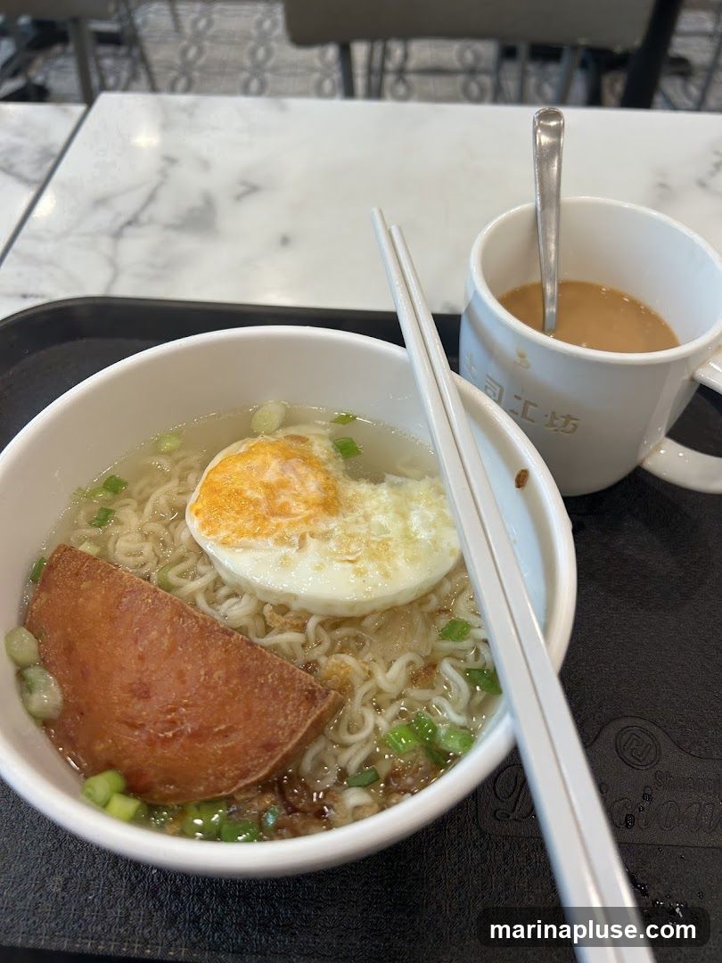 A bowl of instant noodle soup topped with a fried egg and a slice of spam, garnished with chopped green onions. A white mug filled with coffee and a spoon is placed next to the soup. The meal is served on a black tray on a table with a marble-patterned surface.