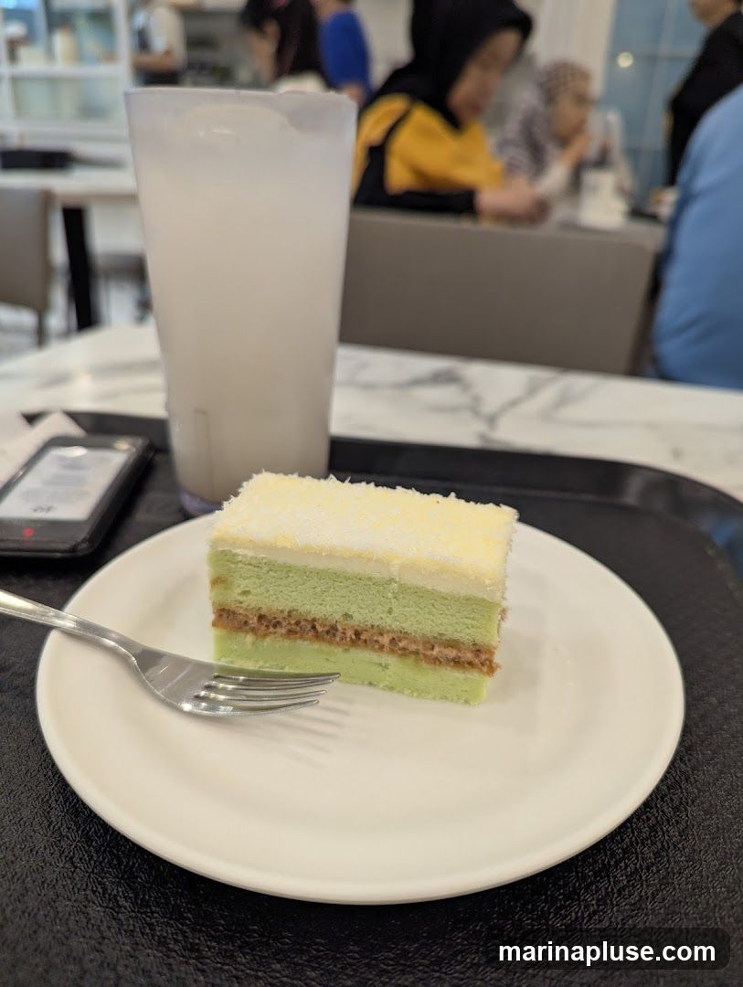 A slice of green cake with white frosting and shredded coconut on top, served on a white plate with a fork. A tall glass of a white beverage with ice is next to the cake. The scene appears to be in a cafe or restaurant with other patrons in the background.