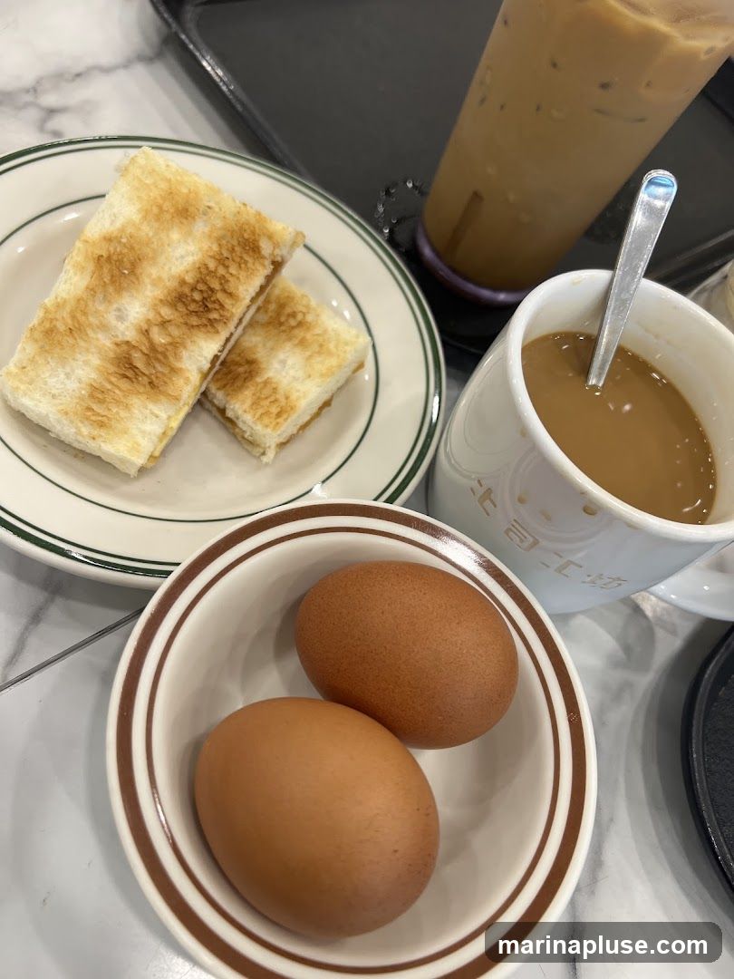 A close-up shot of a breakfast set featuring kaya toast, two soft-boiled eggs, and a cup of coffee. An iced coffee drink is also visible in the background. The toast is golden brown and appears to be buttered. The eggs are whole and unpeeled. The coffee is in a white mug with some text on it.
