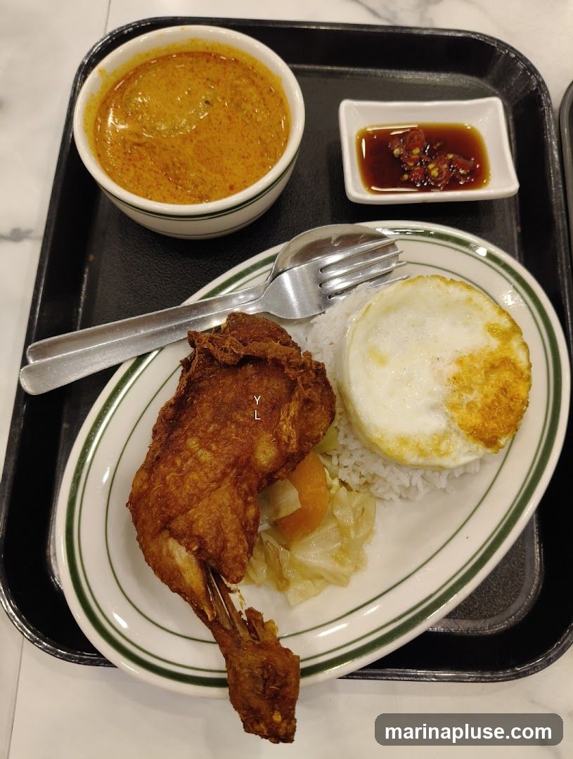A plate of food on a black tray. The plate contains a piece of fried chicken, white rice, a fried egg, and some cooked cabbage and carrots. A small bowl of curry sauce and a small dish of soy sauce with chili flakes are also on the tray.