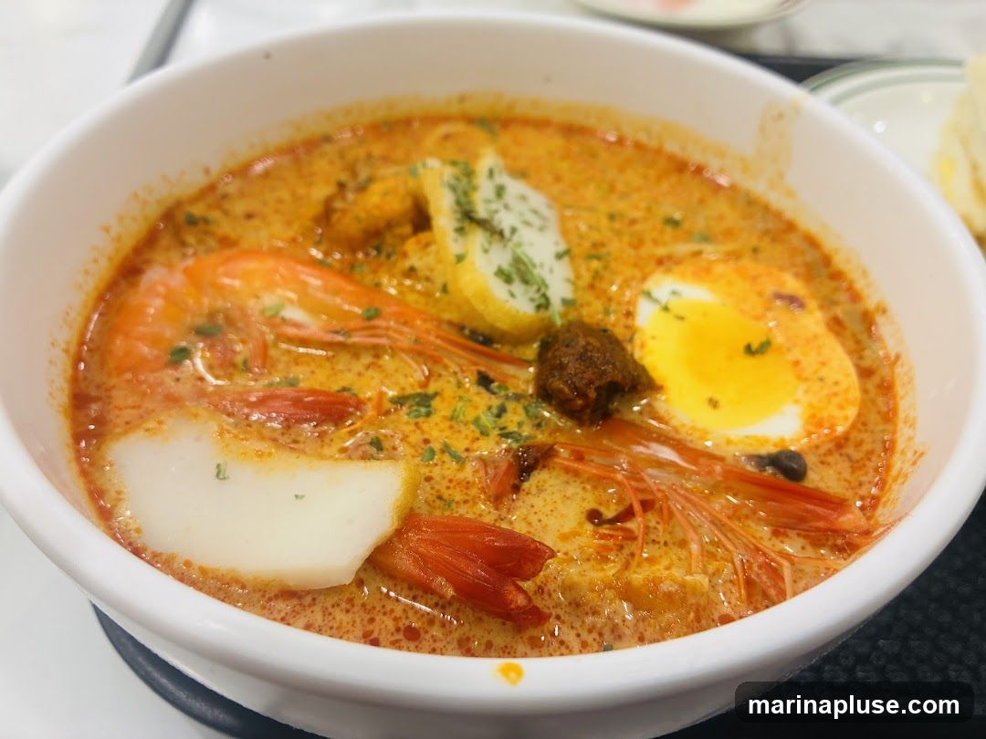 A close-up shot of a bowl of laksa, a spicy noodle soup. The dish features a rich, orange broth, a large shrimp, a halved hard-boiled egg with a runny yolk, slices of fish cake, and a dollop of chili paste. The soup is garnished with chopped herbs.