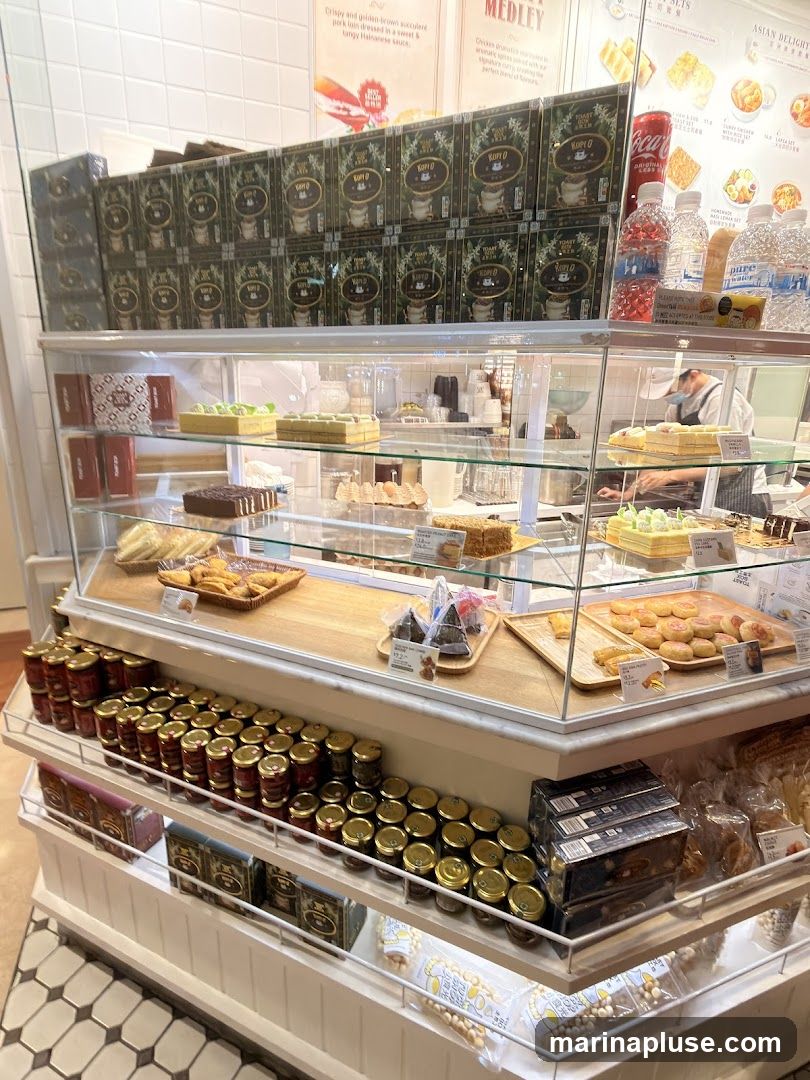 A display case filled with various desserts and packaged goods. On the top shelf, there are boxes of Kopi O coffee. Below, the display case shows an assortment of pastries, cakes, and tarts. The lower shelves are stocked with jars of preserves and packaged snacks. A person in a mask is visible in the background, working behind the counter.