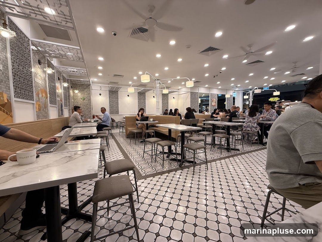 The image shows the interior of a bustling cafe with many tables and chairs occupied by customers. Some individuals are seen working on laptops, while others are engaged in conversations or enjoying their meals. The cafe features a modern design with white walls, patterned tiles on the floor, and contemporary lighting fixtures. The overall atmosphere appears lively and social.