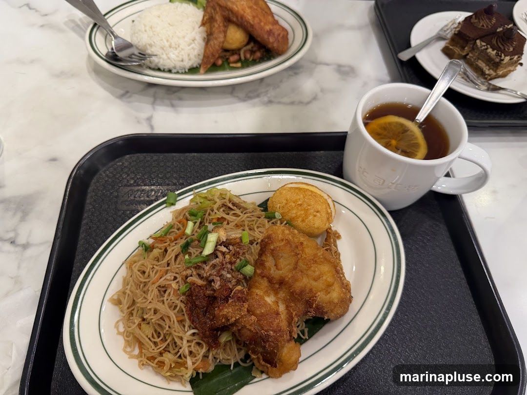 A close-up shot of a meal served on a black tray. The meal includes a plate of fried noodles with fried chicken and a hard-boiled egg, a cup of tea with a lemon slice, and a plate of rice with fried chicken wings and a hard-boiled egg in the background. A slice of chocolate cake is also visible in the upper right corner. The food is presented on white plates with green trim. The mug has some writing on it.