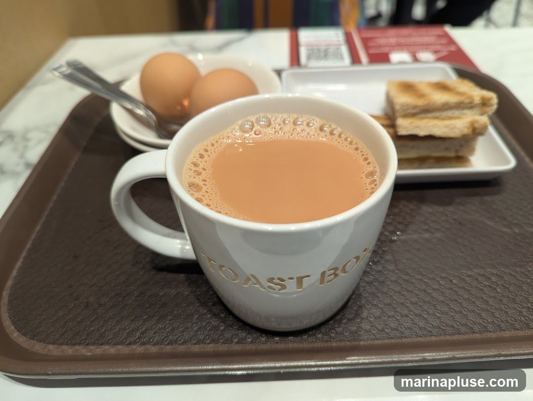 A close-up shot of a breakfast set on a brown tray. In the foreground, a white mug with the embossed text "TOAST BOX" is filled with a frothy, light brown beverage, likely tea or coffee. To the left of the mug, a small white plate holds two soft-boiled eggs and a spoon. In the background, another white plate contains a stack of four slices of toasted bread. The setting appears to be indoors, possibly a cafe or restaurant.