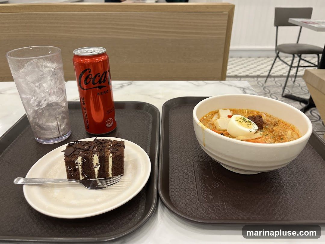 A tray holds a glass of iced water, a can of Coca-Cola Zero, a slice of chocolate cake with white cream filling and chocolate shavings, and a bowl of laksa noodles topped with a hard-boiled egg. The background shows a cafe or restaurant interior with tables and chairs.