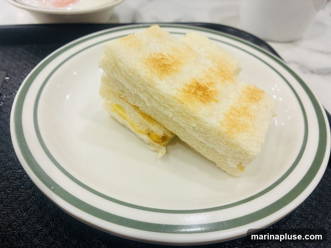 A close-up shot of two slices of toasted bread, cut in half diagonally, revealing a layer of kaya jam and butter filling. The toast is served on a white plate with green stripes. In the background, a soft-focus view of a bowl with what appears to be soft-boiled eggs and a white cup can be seen.