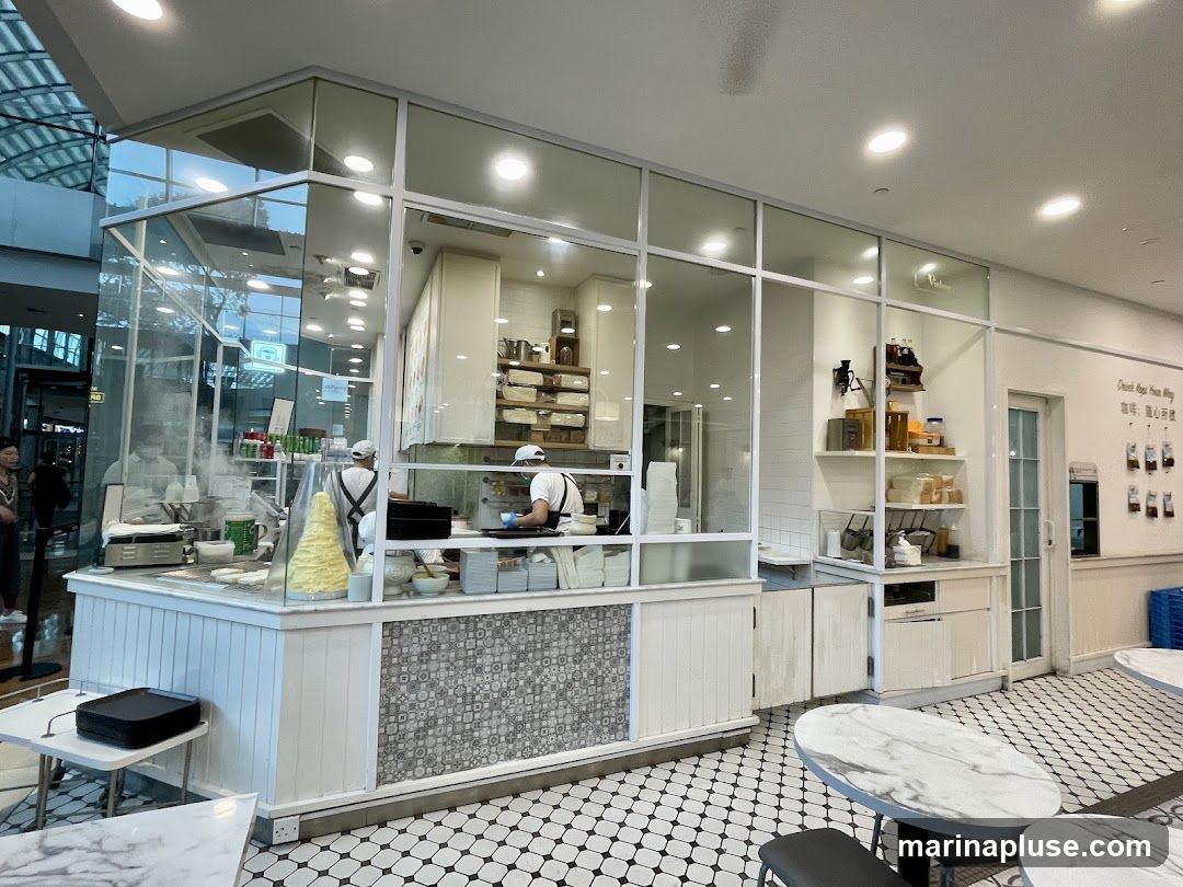 A view of a cafe interior with staff members working behind a counter. The counter area is enclosed by glass, allowing a clear view of the food preparation. One staff member is seen cooking on a griddle, with steam rising. Another staff member is arranging items on a shelf. The cafe has a clean, modern aesthetic with white walls, tiled floors, and marble-topped tables. Shelves are stocked with various ingredients and containers.