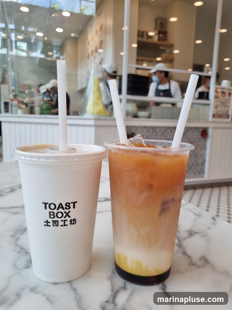 Two drinks are on a marble table. The drink on the left is in a white paper cup with the "TOAST BOX" logo and Chinese characters. The drink on the right is in a clear plastic cup with a layered beverage, possibly tea with milk and syrup. In the background, staff members are visible behind a counter and a clear partition.