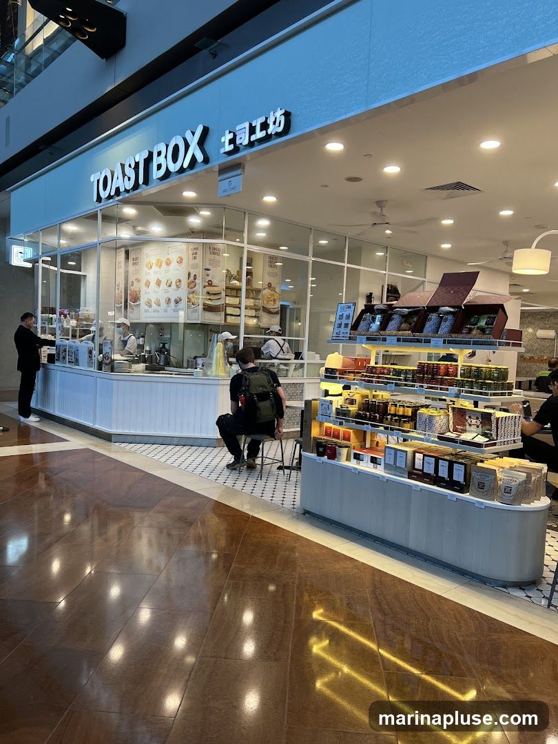 The exterior of a Toast Box cafe with its prominent signage. Inside, staff are visible behind the counter, and customers are seated at tables. Display shelves showcase various food items and packaged goods.