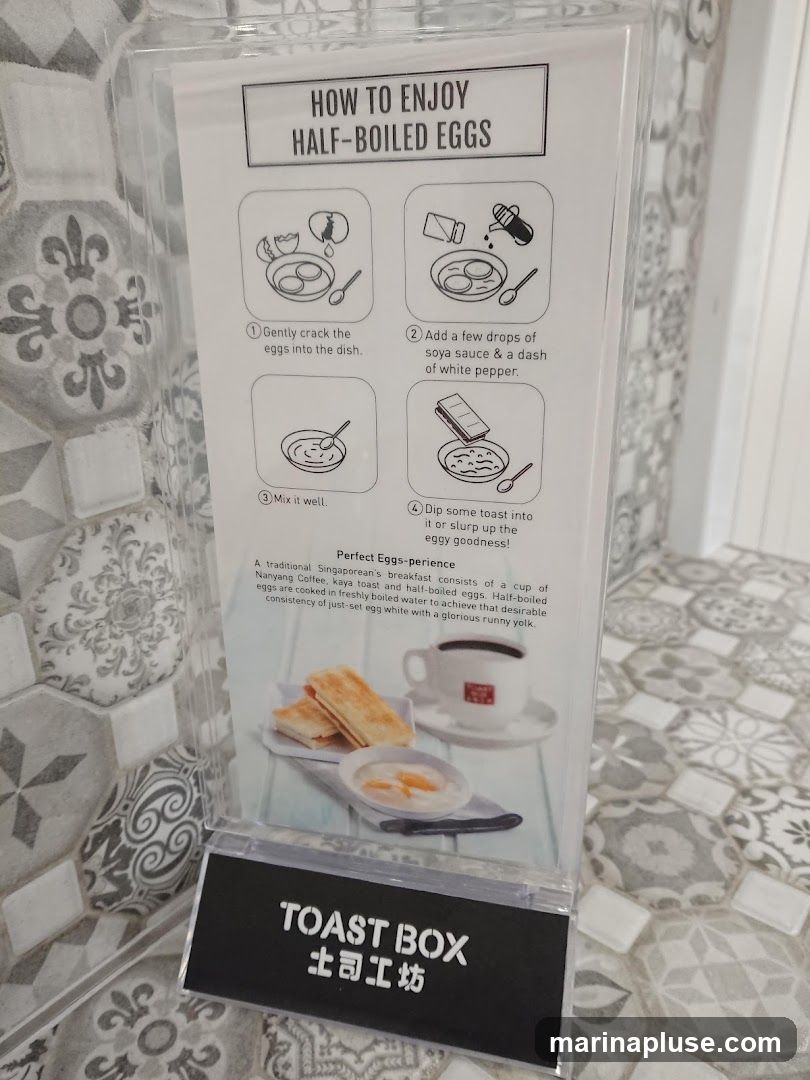 A sign with instructions on how to enjoy half-boiled eggs, including steps like cracking eggs, adding soy sauce and pepper, mixing, and dipping toast. It also describes the perfect eggs-perience of a traditional Singaporean breakfast with Nanyang Coffee and kaya toast. The sign has the 'TOAST BOX' logo at the bottom.