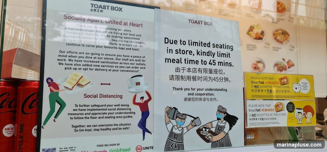 Signs at a Toast Box store display information regarding social distancing, limited seating, and the non-acceptance of BreadTalk Rewards. The signs are in English and Chinese. Also visible are cans of Coca-Cola Zero.