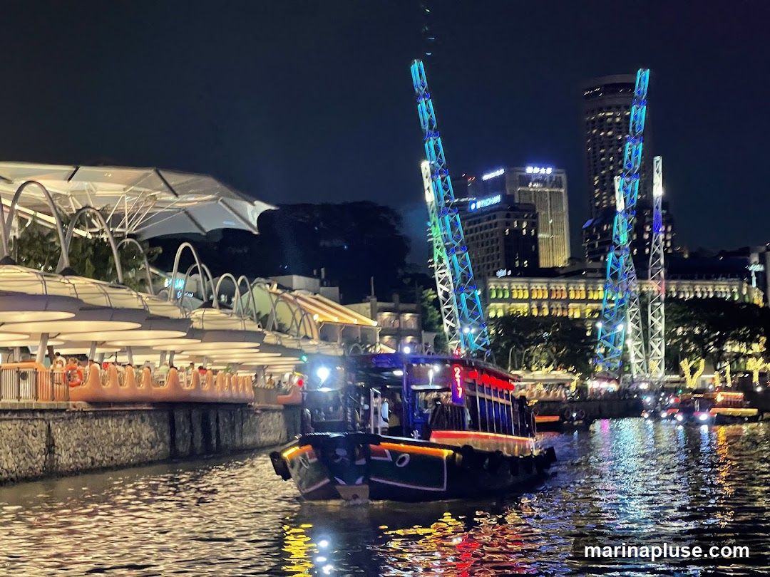 A boat with colorful lights cruises down a river at night. In the background, a cityscape with illuminated buildings and tall, brightly lit towers is visible. Along the riverbank, there are covered seating areas with lights and people.