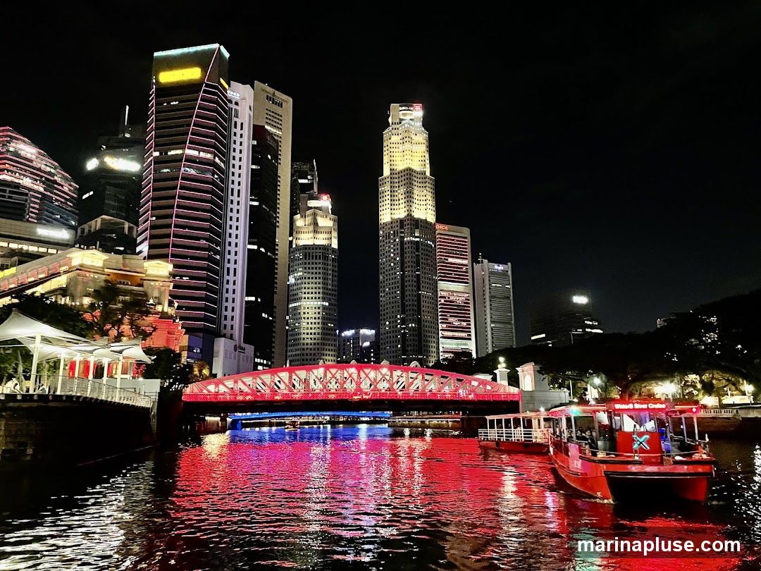 A nighttime view of a city skyline featuring illuminated skyscrapers and a river. A red bridge spans the river, reflecting red and blue lights onto the water. Two red boats are on the river, one with "Water B River Cruise" visible on its side. The buildings are lit up with various colors, creating a vibrant urban scene.