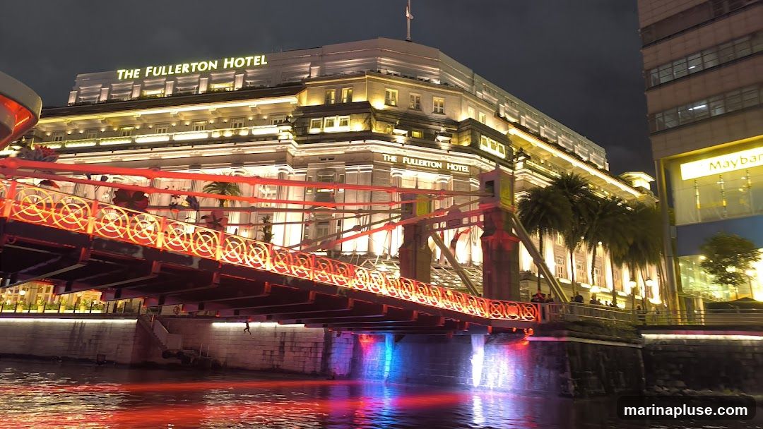 A nighttime view of The Fullerton Hotel, a grand building illuminated with warm lights. A bridge with intricate red and orange lighting spans across a body of water in the foreground, reflecting the colorful lights. Palm trees are visible along the waterfront. To the right, a modern building with a yellow sign that reads "Maybank" is partially visible.