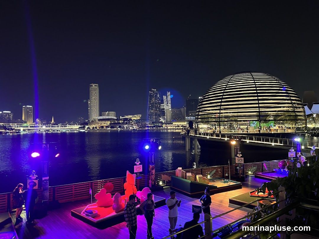 A nighttime view of a waterfront area with a distinctive domed building in the background. In the foreground, there is a mini-golf course with colorful obstacles and people playing. The area is illuminated with purple and pink lights, and a cityscape is visible across the water.