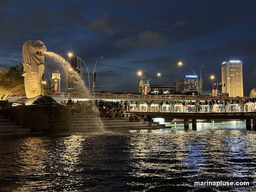 A wide shot of the Merlion fountain in Singapore at night. The iconic statue is spouting water into the Singapore River. Many people are gathered on the steps and a bridge overlooking the fountain. The city skyline is visible in the background, with illuminated buildings and streetlights reflecting on the water.