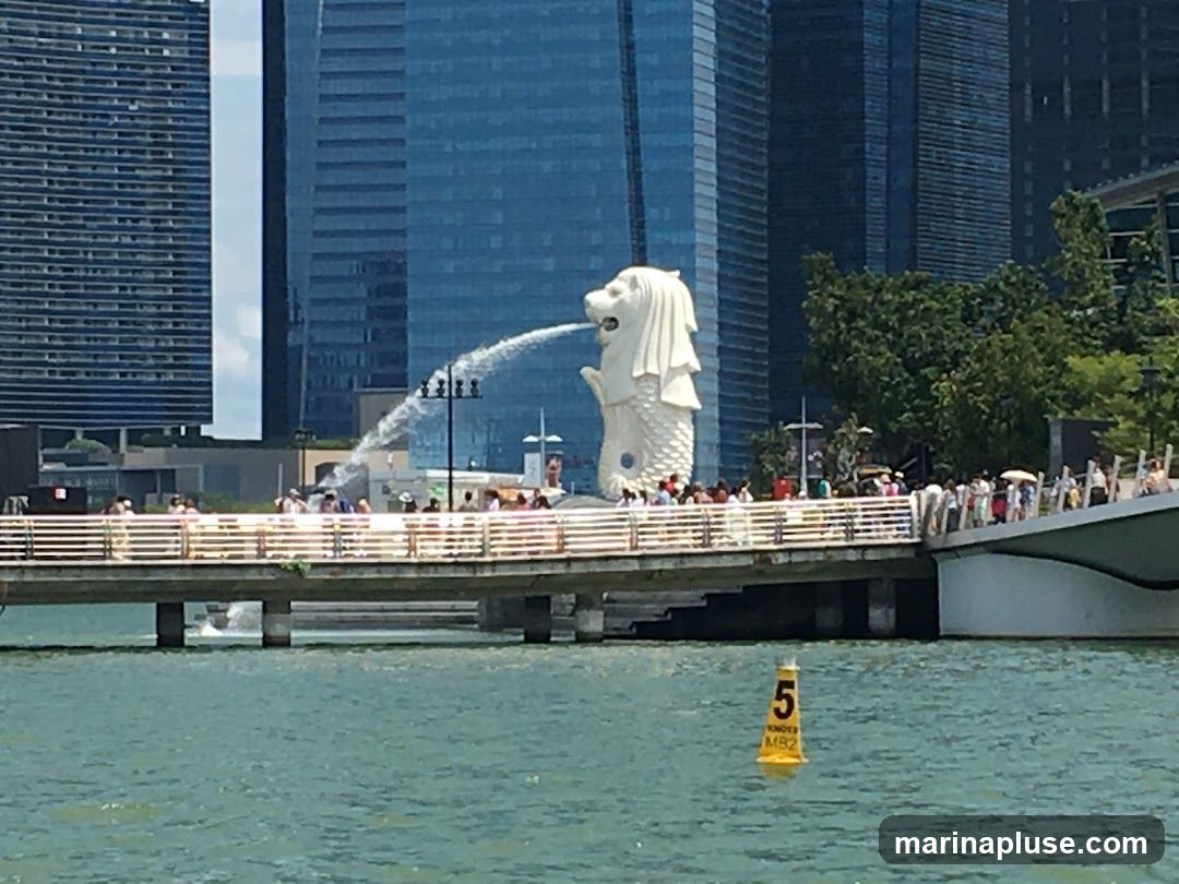 A view of the Merlion statue in Singapore, with its iconic water spout, set against a backdrop of modern skyscrapers. A pedestrian bridge filled with people spans across the water in the foreground, leading towards the statue. The water is a vibrant turquoise, and a yellow buoy with the number '5' is visible in the lower right corner.