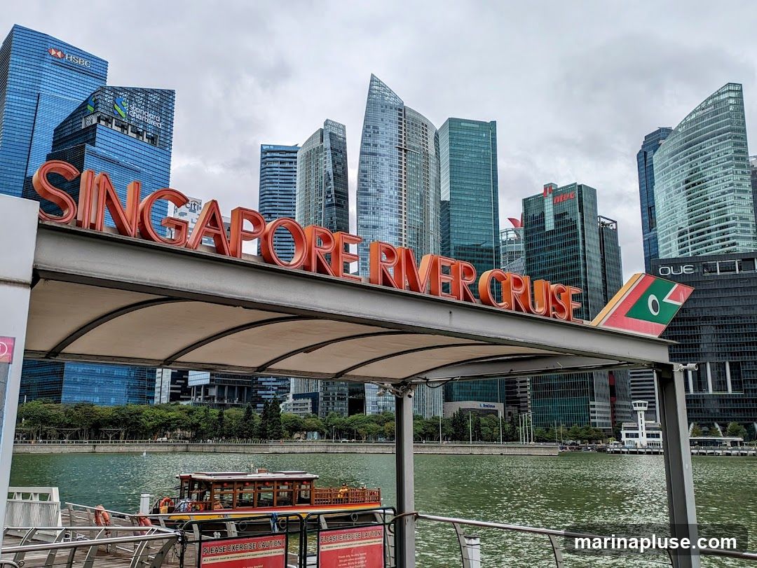 A large, orange, three-dimensional sign spelling out "SINGAPORE RIVER CRUISE" is prominently displayed against a backdrop of modern skyscrapers and a cloudy sky. The sign is mounted on a covered walkway structure. To the right of the sign, a stylized airplane wing graphic with green and red accents is visible. Below the walkway, a river is visible with a boat docked at a pier.