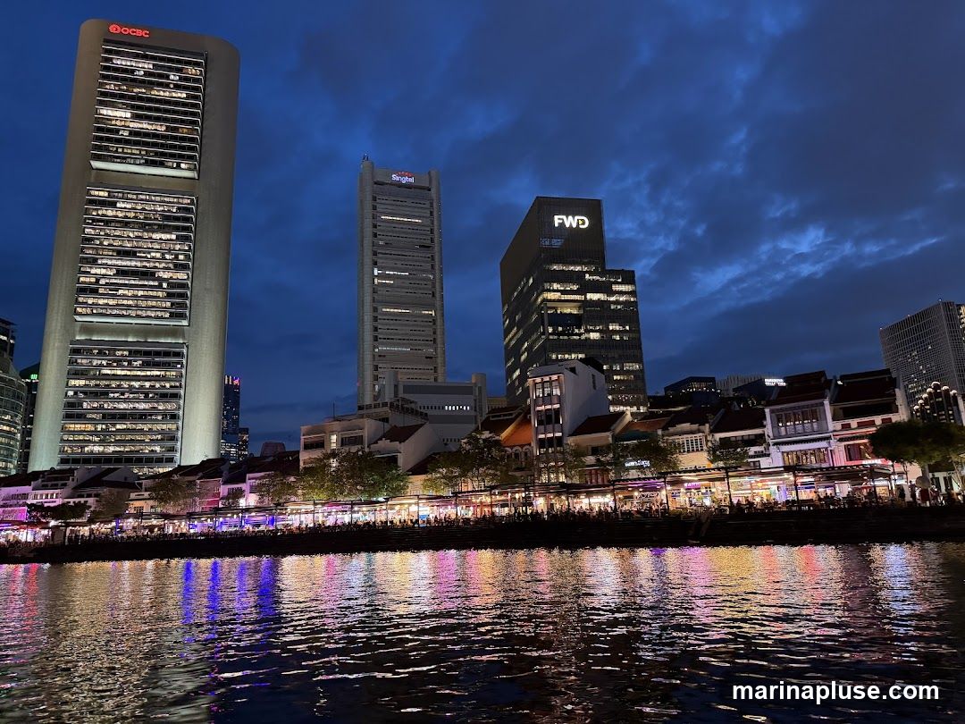 A nighttime view of a city skyline featuring several tall office buildings, including one with the OCBC logo and another with the Singtel logo. Below the skyscrapers, a row of lower buildings with illuminated storefronts lines a waterfront. The lights from the buildings and storefronts are reflected in the dark water of a river in the foreground, creating a colorful and shimmering effect. The sky is a deep blue with dramatic clouds.