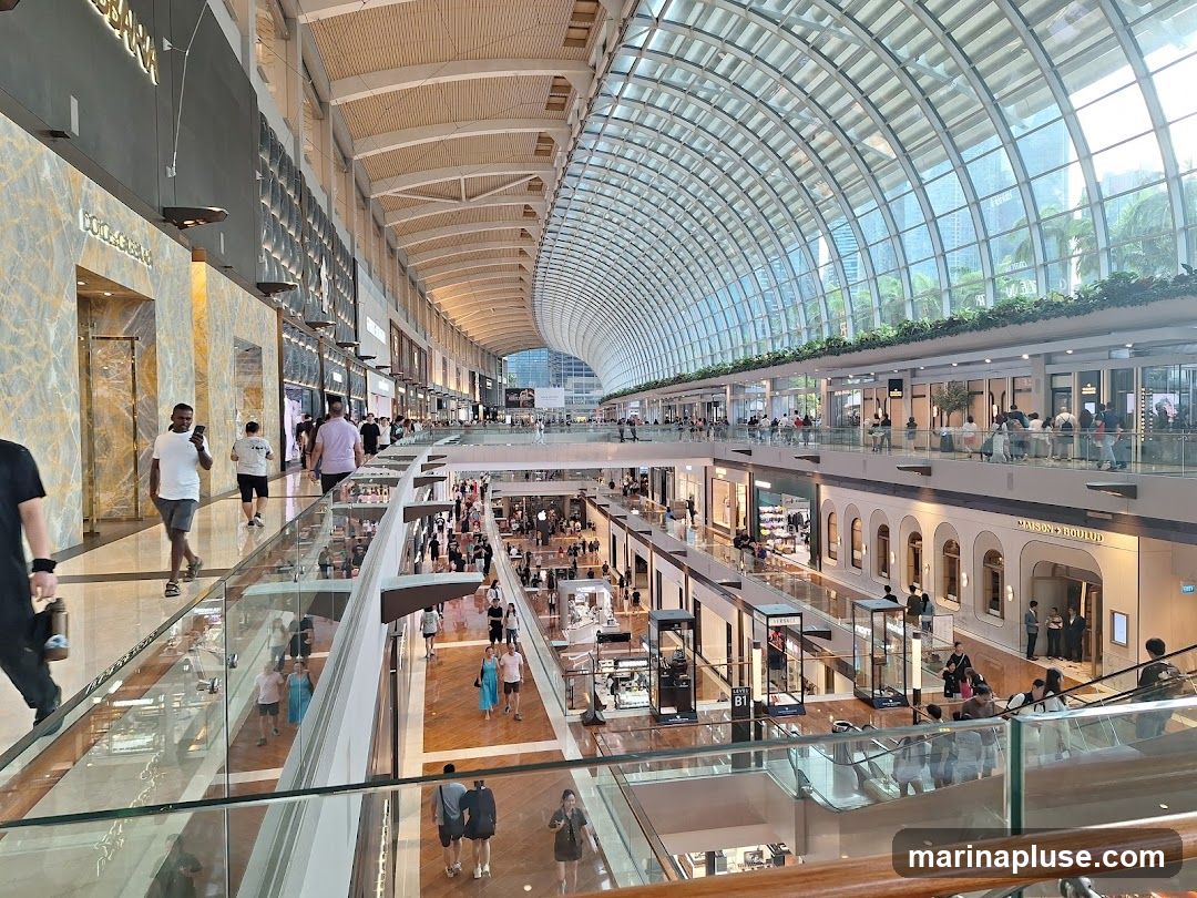 A wide-angle view of the interior of a multi-level shopping mall with a large glass-curved roof. People are seen walking on the various levels, browsing shops, and using escalators. The shops have modern facades, some with marble accents and prominent signage. The overall atmosphere is bright and spacious, with a mix of luxury and retail stores.