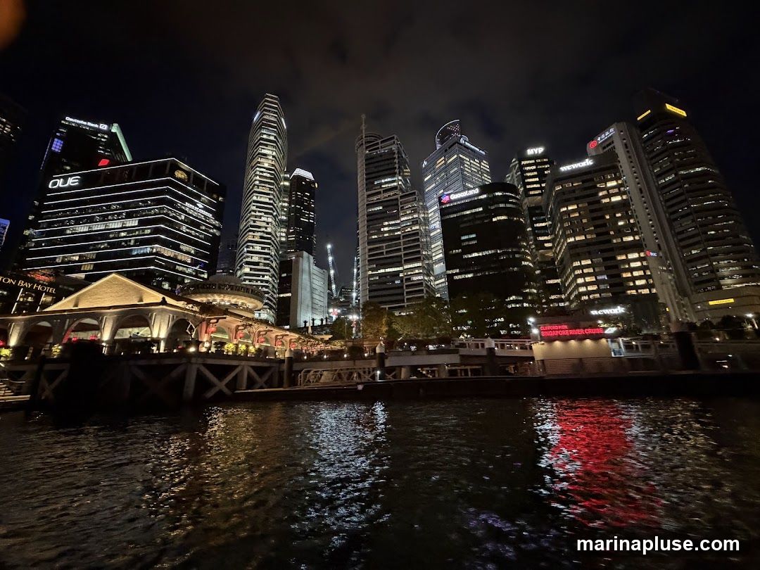 A nighttime view of the Singapore skyline, featuring illuminated skyscrapers and a waterfront building reflected in the dark water. The buildings display various corporate logos and architectural designs.