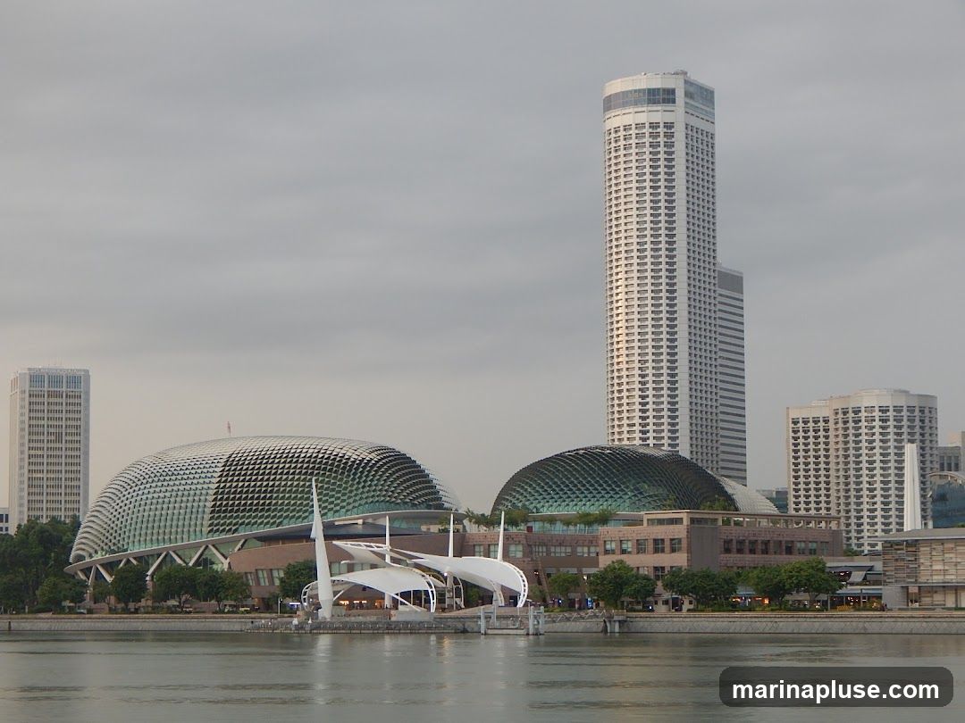 A wide shot of the Singapore skyline featuring the iconic Marina Bay Sands hotel and the Esplanade Theatres on the Bay, also known as the durian. The buildings are situated along the waterfront, with a body of water in the foreground. The sky is overcast, casting a soft light on the scene. Several other skyscrapers are visible in the background, contributing to the dense urban landscape.