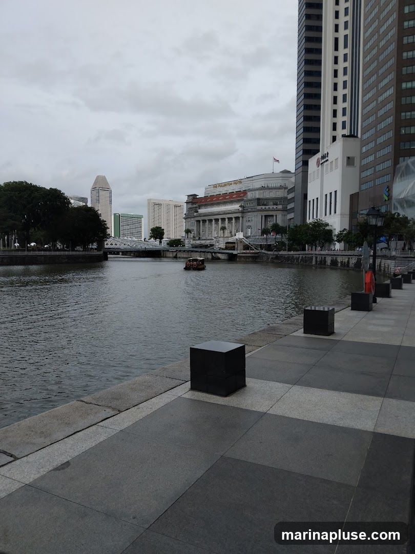 A view of the Singapore River with buildings and a boat. The Fullerton Hotel is visible on the right side of the river. The walkway in the foreground has a checkered pattern of dark and light gray tiles, with black cube-shaped bollards. The sky is overcast.