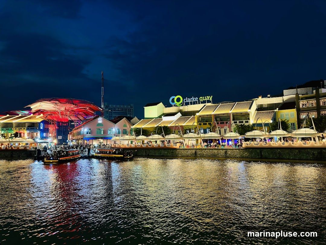 A nighttime view of Clarke Quay, a riverside area with colorful buildings and outdoor seating. Boats are docked along the water, and the area is illuminated by lights, reflecting on the water. The sign for 'clarke quay' is visible on one of the buildings.