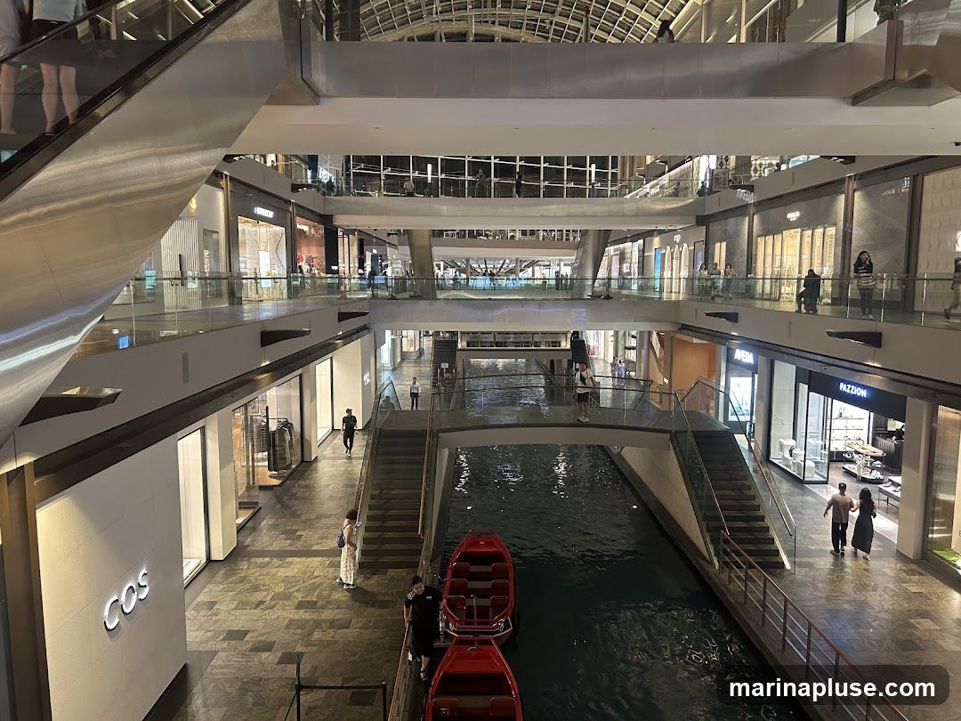 An interior view of a multi-level shopping mall featuring a canal with gondola-like boats. Shops with illuminated storefronts line the walkways on different levels. Escalators and staircases connect the floors, and glass railings overlook the canal and lower levels. People are seen walking and browsing.