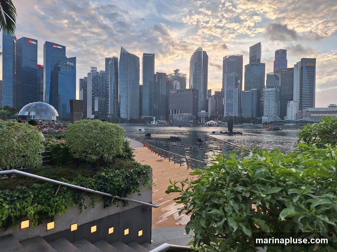 A panoramic view of the Singapore skyline at dusk, with modern skyscrapers dominating the background. In the foreground, lush green bushes and trees frame a walkway along the water. The sky is filled with dramatic clouds, illuminated by the setting sun. Several boats are visible on the water, and a distinctive domed building is present on the left side of the image.