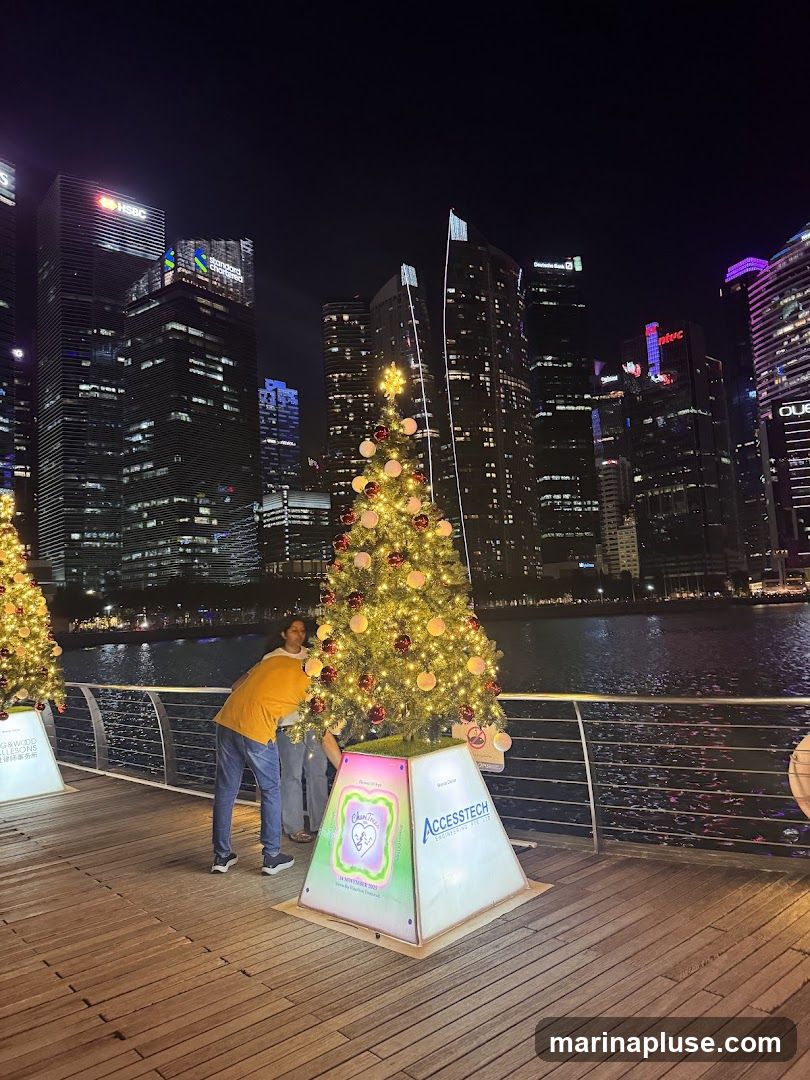 Two decorated Christmas trees stand on a wooden boardwalk at night. In the background, a city skyline with illuminated skyscrapers is visible across a body of water. One tree has a sign at its base with the text "AccessTech" and "ENGINEERING THE BITS". Another sign on a tree further left has Chinese characters and the text "CHRISTMAS".