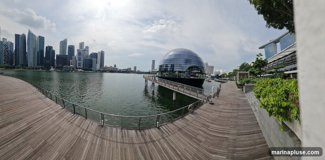 A panoramic view of a modern waterfront promenade with a distinctive domed building on the water. The promenade is made of wooden planks and has metal railings. In the background, a dense city skyline with numerous skyscrapers is visible across the water. The domed building, which appears to be a restaurant or entertainment venue, is connected to the promenade by a walkway. Lush greenery lines parts of the promenade, and the sky is partly cloudy.