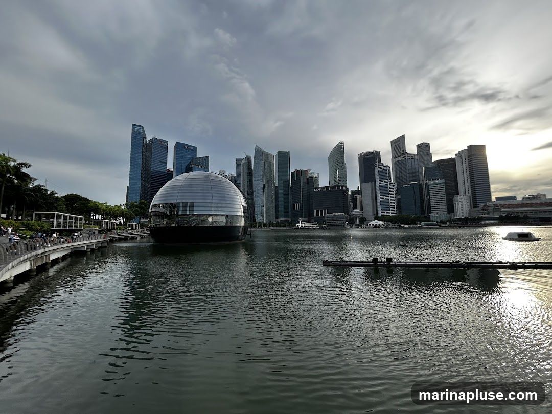 A wide shot of the Singapore skyline at dusk, featuring the iconic Marina Bay Sands hotel and the ArtScience Museum, which resembles a lotus flower. The buildings are reflected in the calm waters of Marina Bay. A walkway with people can be seen on the left. The sky is cloudy with the sun setting on the right.