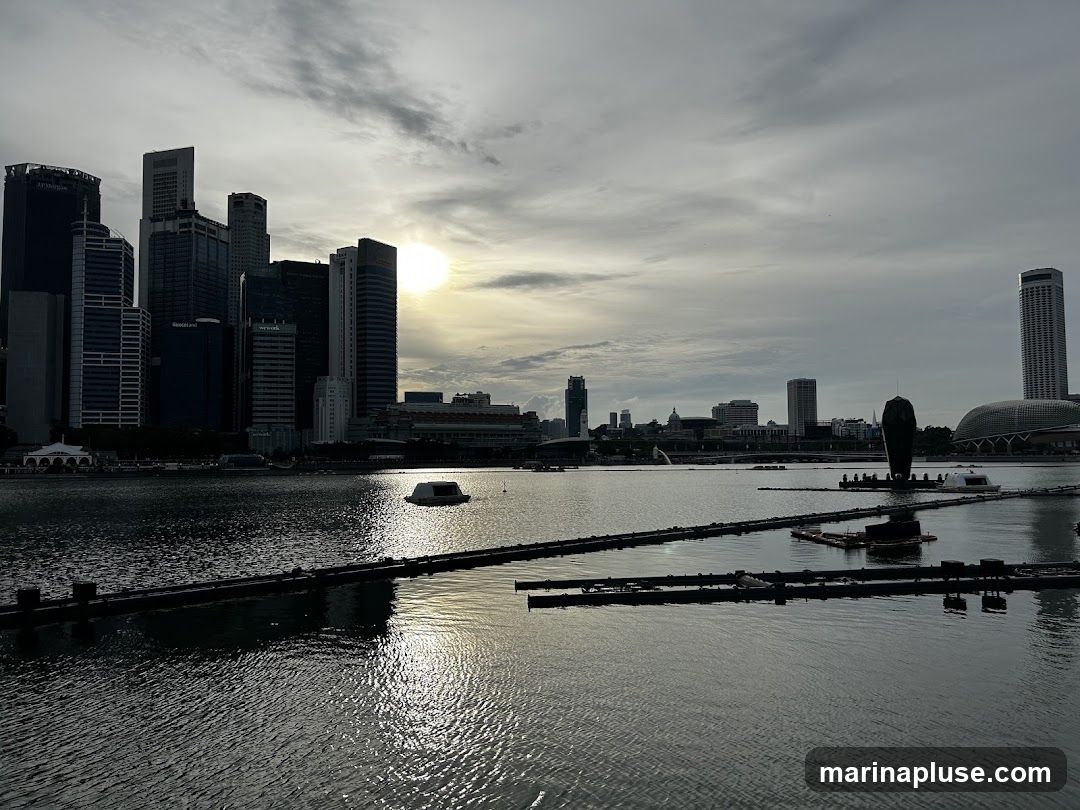A panoramic view of the Singapore skyline at dusk, with tall skyscrapers silhouetted against a cloudy sky. The sun is setting, casting a warm glow on the horizon. The water in the foreground reflects the city lights and the sky. Several floating structures and a large, dark sculpture are visible in the bay.