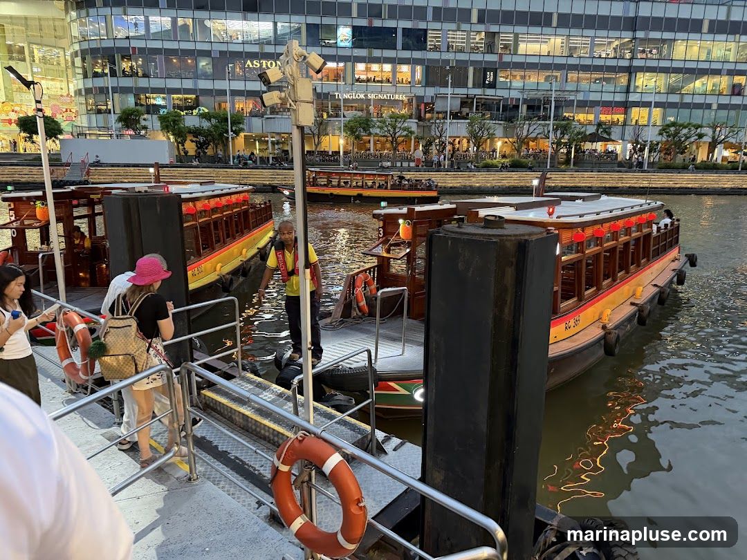 People are boarding or disembarking from traditional river boats docked at a pier. The boats are yellow and orange with red lanterns. In the background, a modern glass-fronted building with multiple stories is visible, featuring various shops and restaurants. The water reflects the lights from the building and the boats.