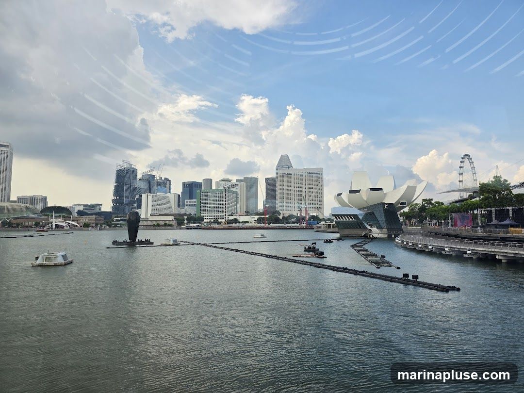 A wide shot of the Singapore skyline, featuring Marina Bay with its iconic buildings and structures. In the foreground, the water of the bay is calm. In the mid-ground, the ArtScience Museum, shaped like a lotus flower, is prominent. To its right, a Ferris wheel is visible. The background shows a dense collection of skyscrapers, representing the city's financial district. The sky is partly cloudy with a mix of blue sky and white clouds. There are also some floating structures and barriers in the water.